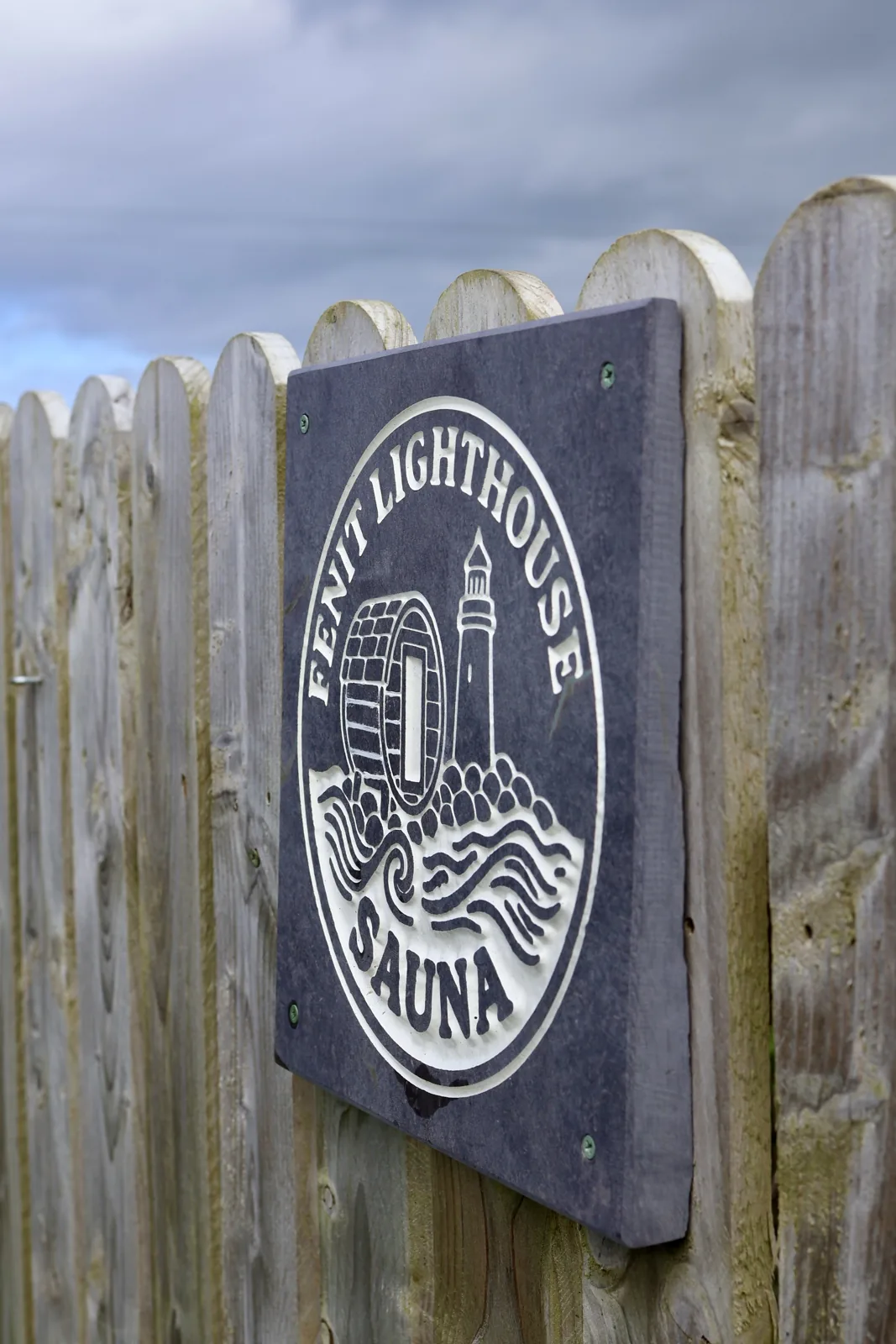 Fenit Lighthouse Sauna sign with its lighthouse and waves emblem on the fence at the entrance