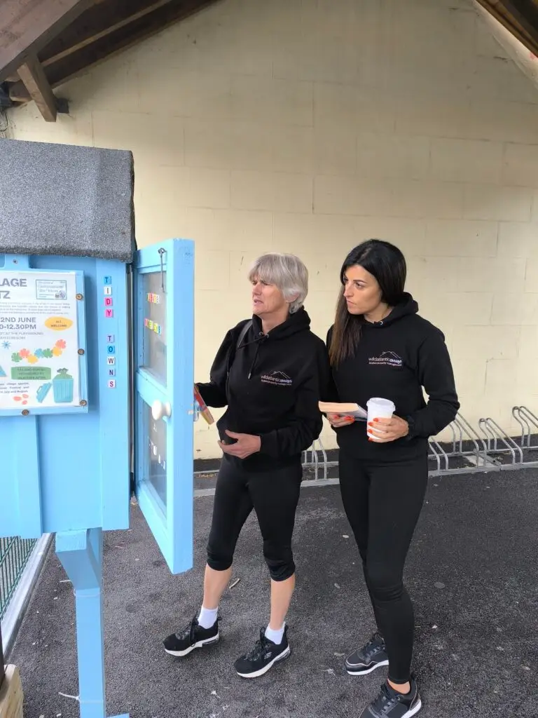 Two women at a book borrow box in Castlegregory. The blue box has a notice and a door. The women wear black tracksuits and trainers. Outdoor setting with bike racks visible.