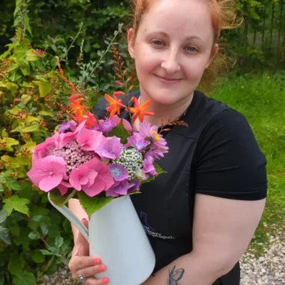 Head housekeeper on the Wild Atlantic Way holding a jug of pink and purple flowers. The housekeeper has red hair and butterfly tattoos on her arm.